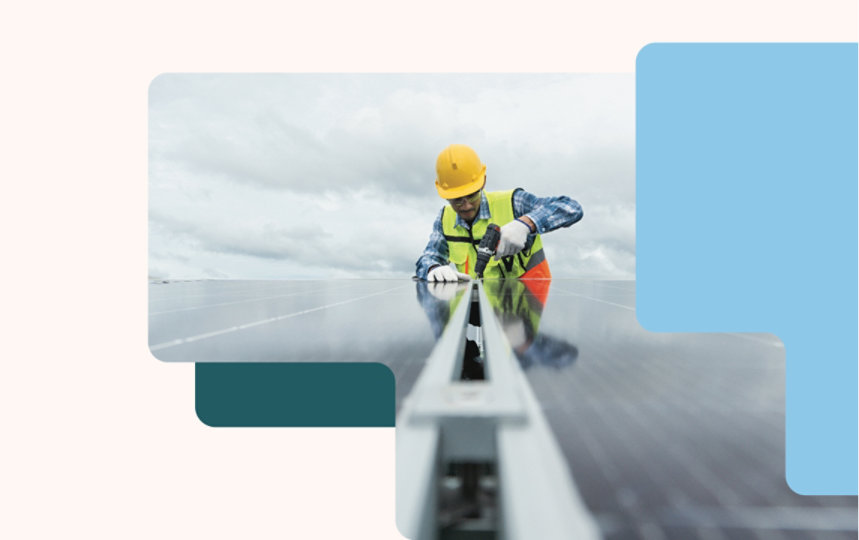 A worker in a hard hat and safety vest installs a solar panel under a cloudy sky.