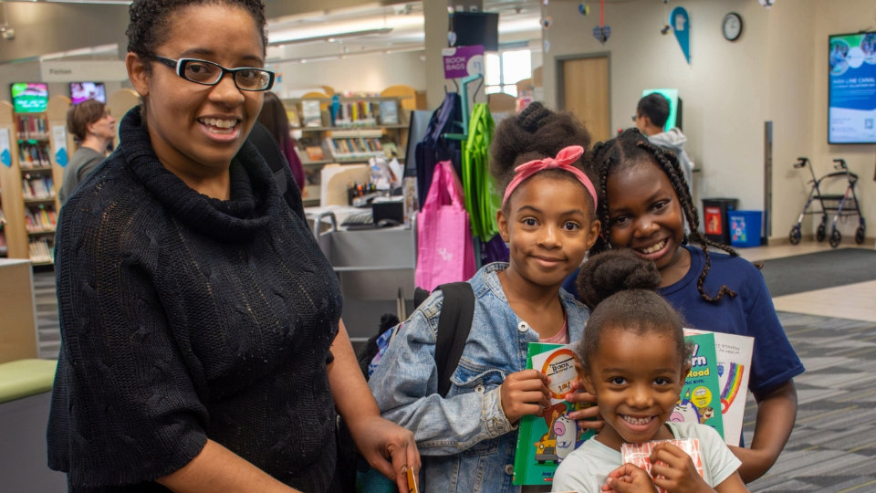 A woman and three smiling children holding books in a library setting.