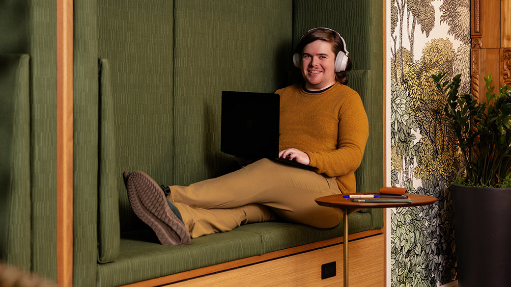 A man sitting on a bench with headphones and a laptop, smiling at the camera in a space filled with natural tones of green, brown and red with warm wood accents.