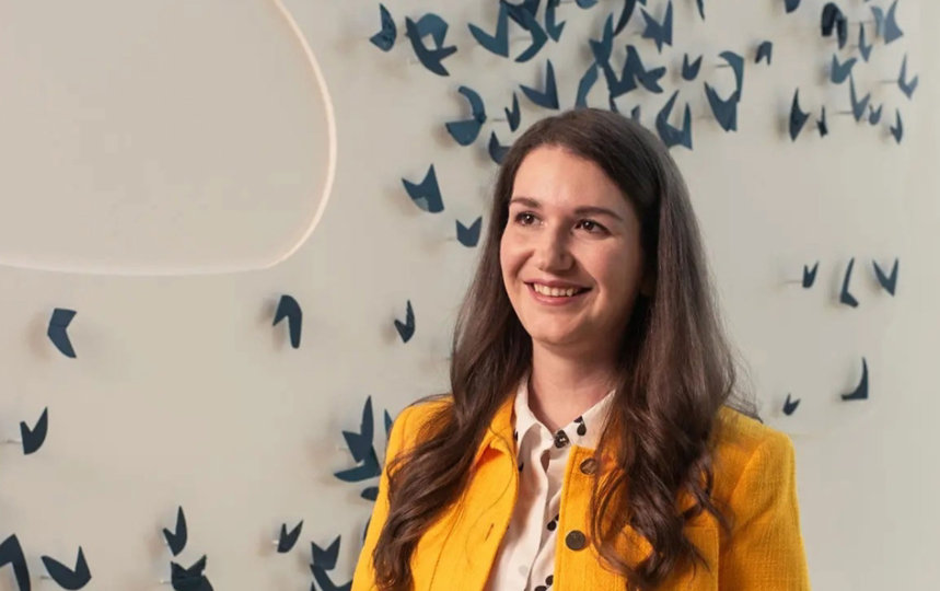Natalie Worden-Cowe, wearing a yellow blazer with a white blouse underneath, standing in front of a white wall with navy decorative art scattered, smiling