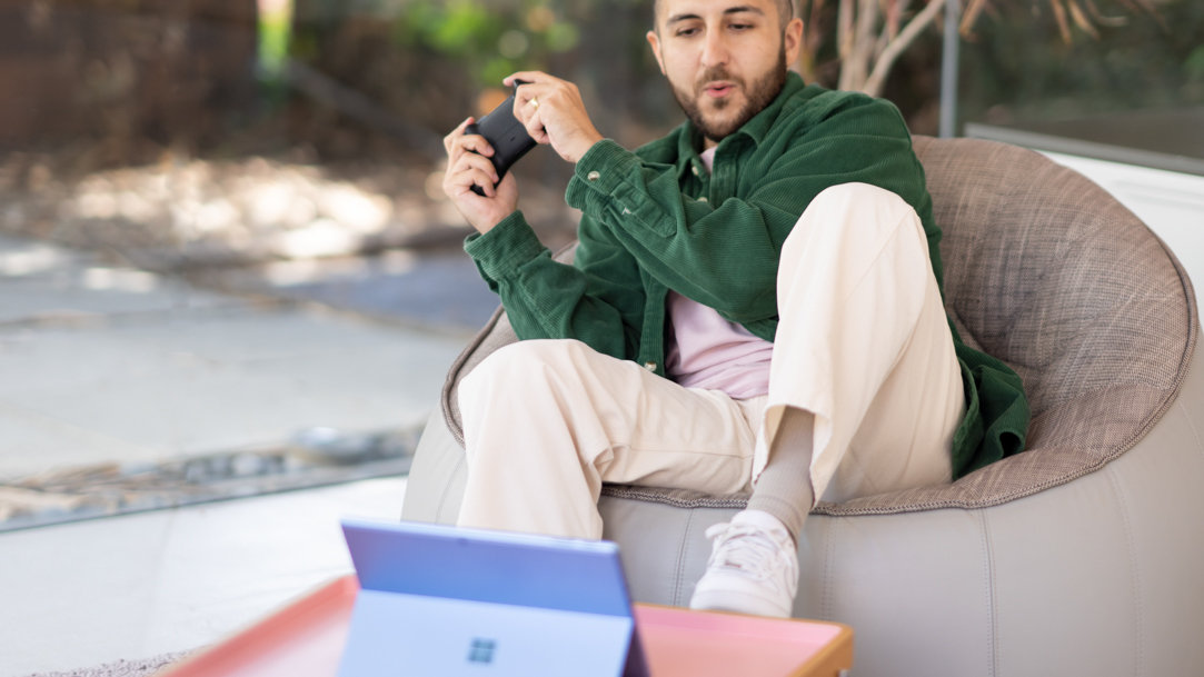A man sitting outside while playing video games using a  controller and a 2-in-1 Surface PC