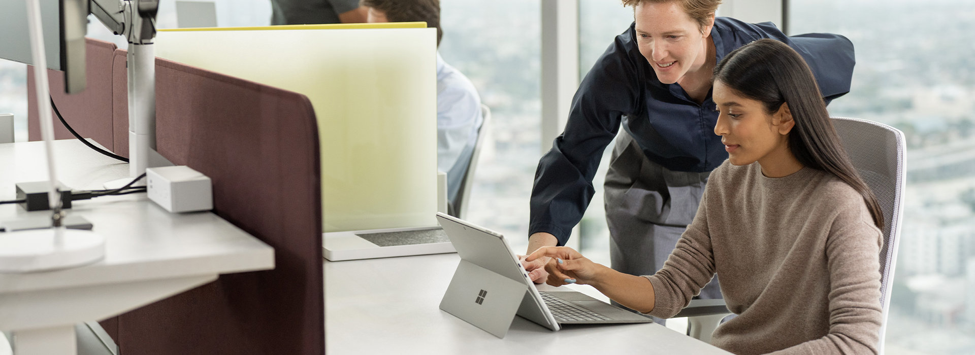 A person looks over the shoulder of someone sitting at an office desk and interacts with the screen of a Surface device
