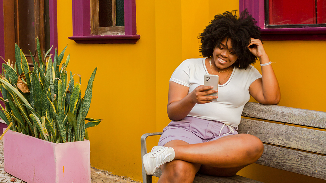 A young woman sitting on a bench in front of a brightly colored building looking at her phone