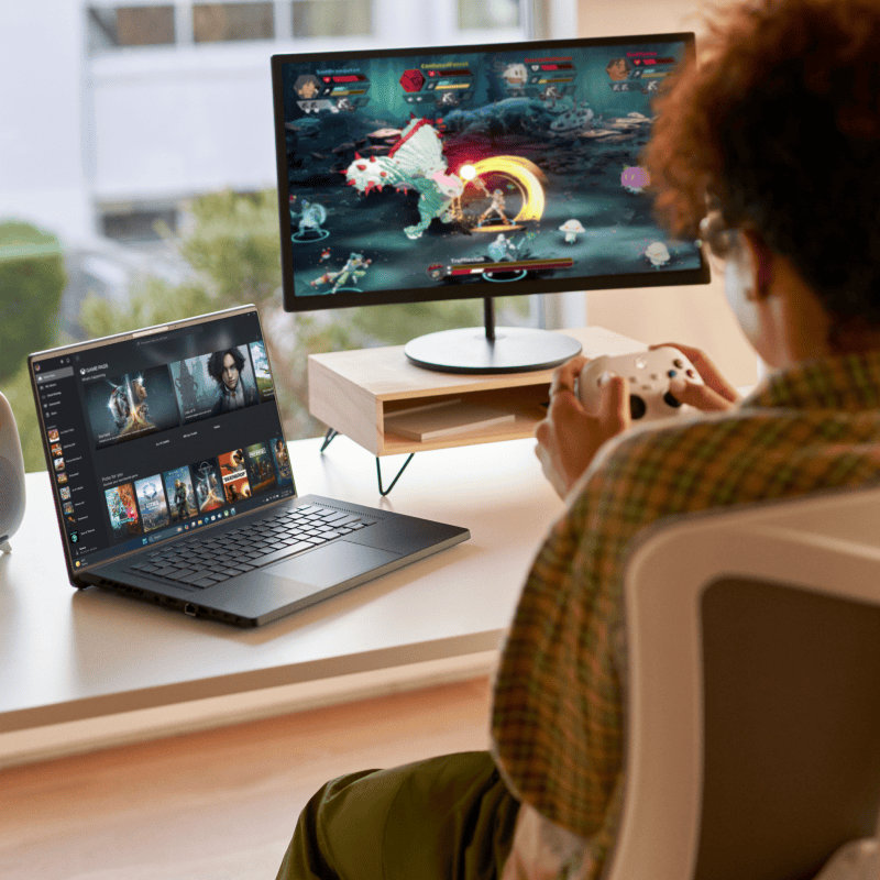 A person sitting at a desk, holding a video game controller, playing a video game on an open laptop and computer monitor