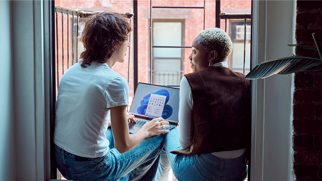 Two Gen Z friends sitting on fire escape balcony with laptop open with Windows 11 start screen and start menu