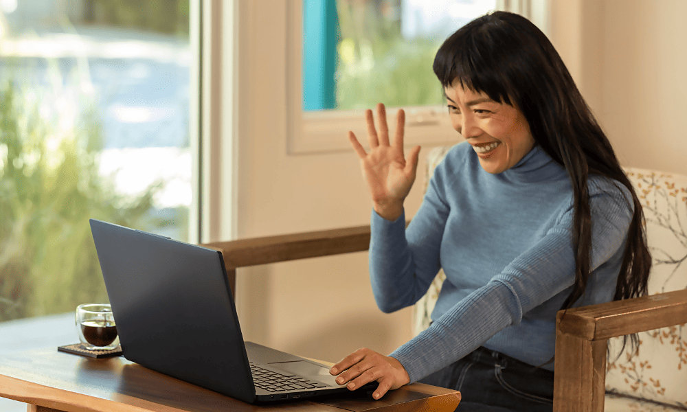 A person smiling and waving at an open laptop while sitting at a desk