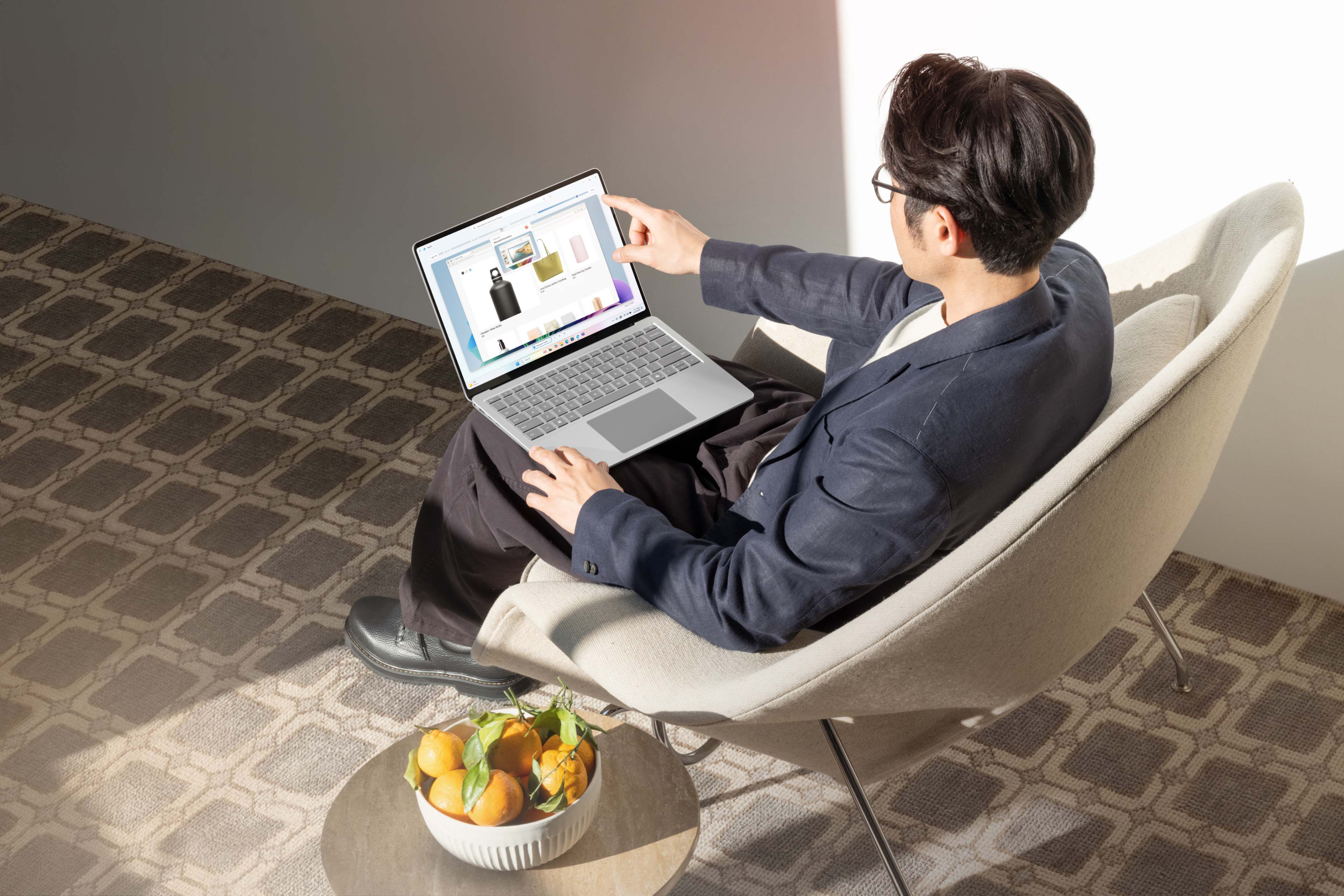 With his back to the viewer, a man sits in a chair using the touchscreen functionality of a Surface Pro to shop online.
