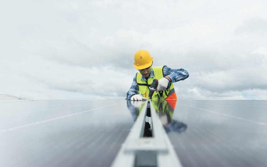 A worker in a hard hat and safety vest installs a solar panel under a cloudy sky.